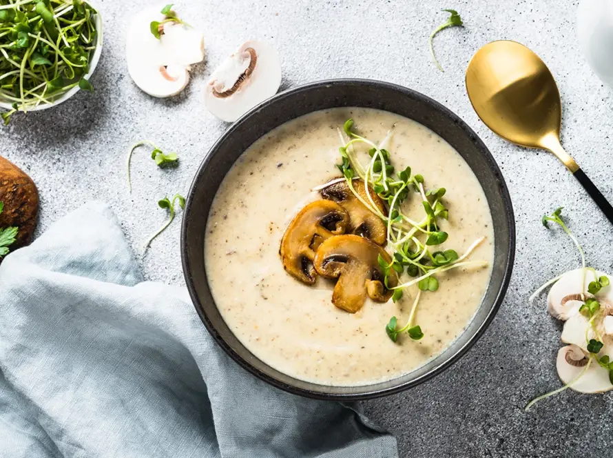 Top view of creamy Champignon mushroom soup in a bowl on a light stone table, featuring a rich texture and fresh herb garnish for B2B food service.